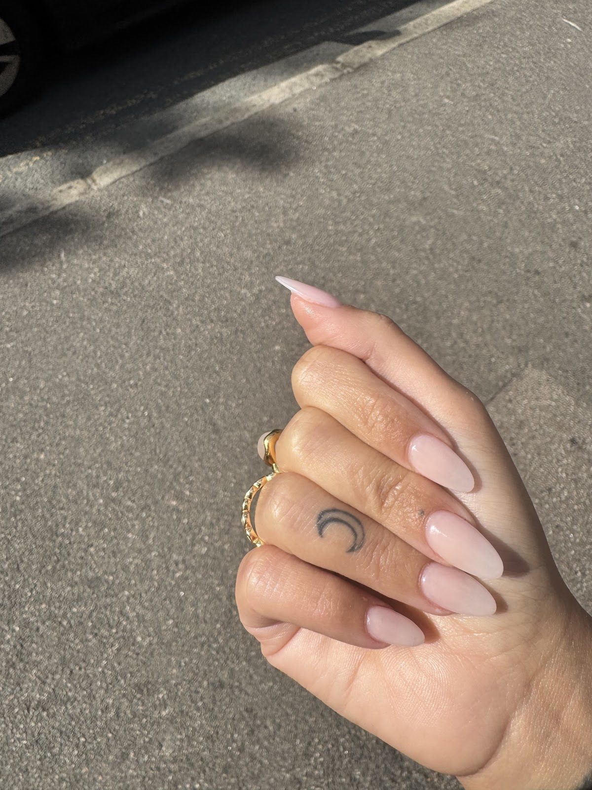 Soft nude almond nails with stacked gold rings, hand held over grey tarmac